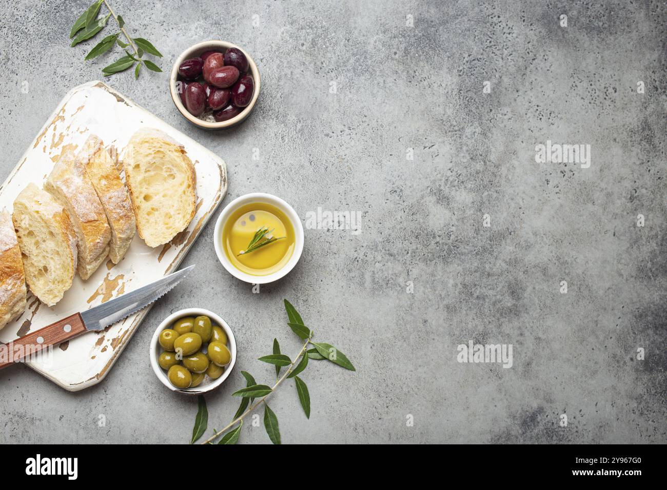 In Scheiben geschnittene frische Ciabatta auf Schneidebrett, grüne und braune Oliven, Olivenöl mit Rosmarin, Olivenzweige auf grauem Betonstein rustikale bac Stockfoto