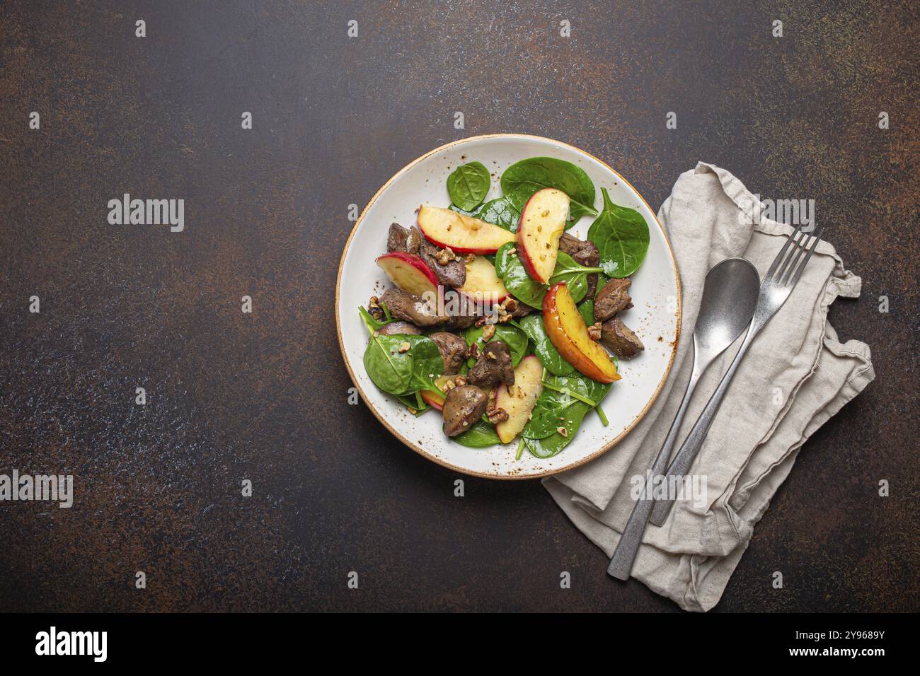 Gesunder Salat mit eisenreichen Zutaten Hühnerleber, Äpfel, frischer Spinat und Walnüsse auf weißer Keramikplatte, dunkelbrauner rustikaler Steinhintergrund T Stockfoto