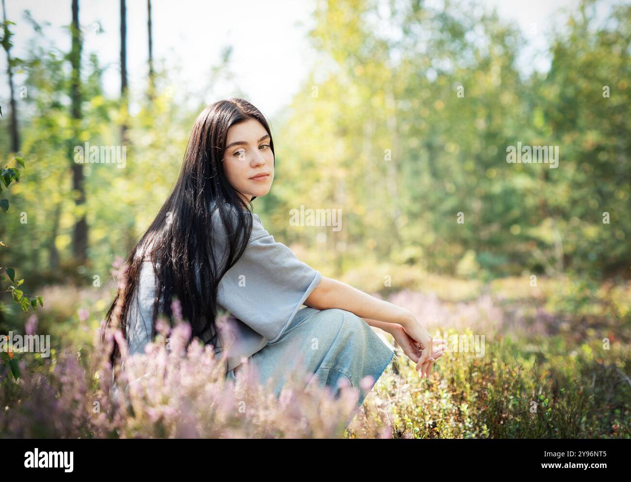 Eine junge Frau sitzt ruhig auf dem Waldboden, ihr langes Haar zieht sich über den Rücken. Sie erscheint tief in Gedanken, umgeben von blühenden Wildblumen Stockfoto