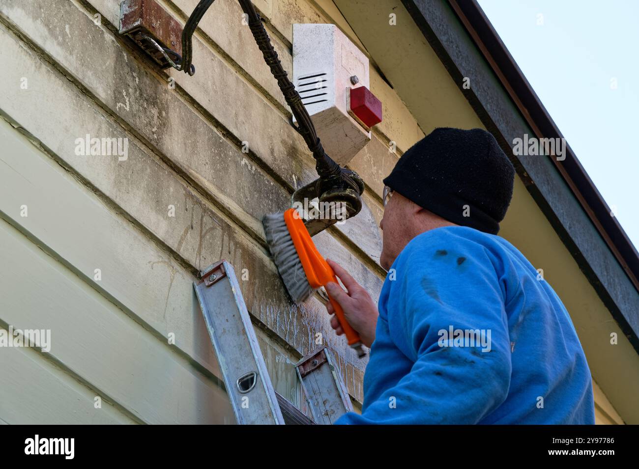 Ein Mann schrubbt ein Haus, bevor er malt. Er ist in der Nähe des elektrischen Eingangs zum Haus und einer Alarmbox Stockfoto