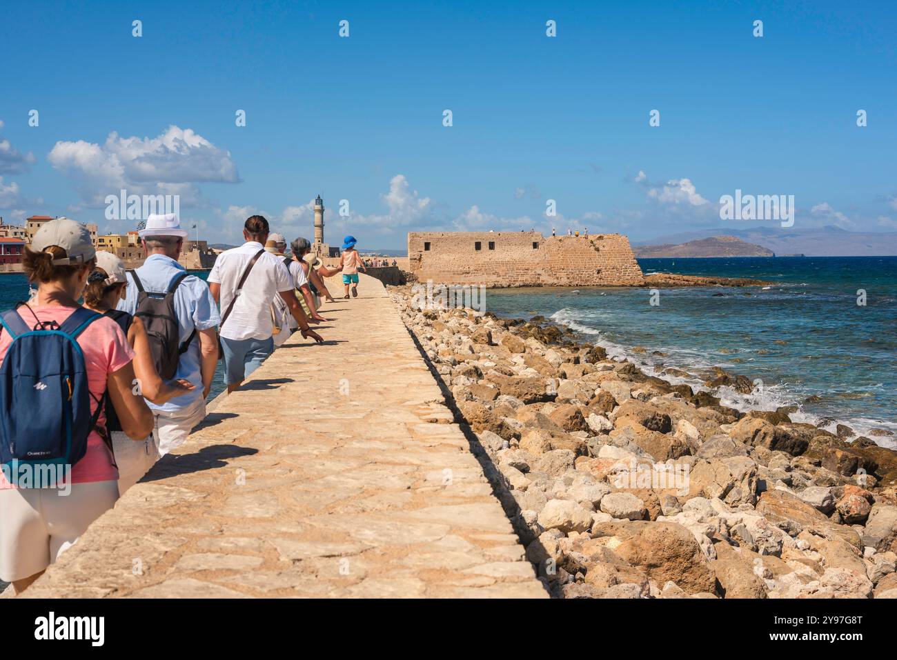 Tourismus Europa, Rückansicht einer Gruppe von Touristen in Chania, Kreta, die in einer einzigen Datei auf der alten venezianischen Hafenmauer in Richtung eines Leuchtturms laufen. Stockfoto