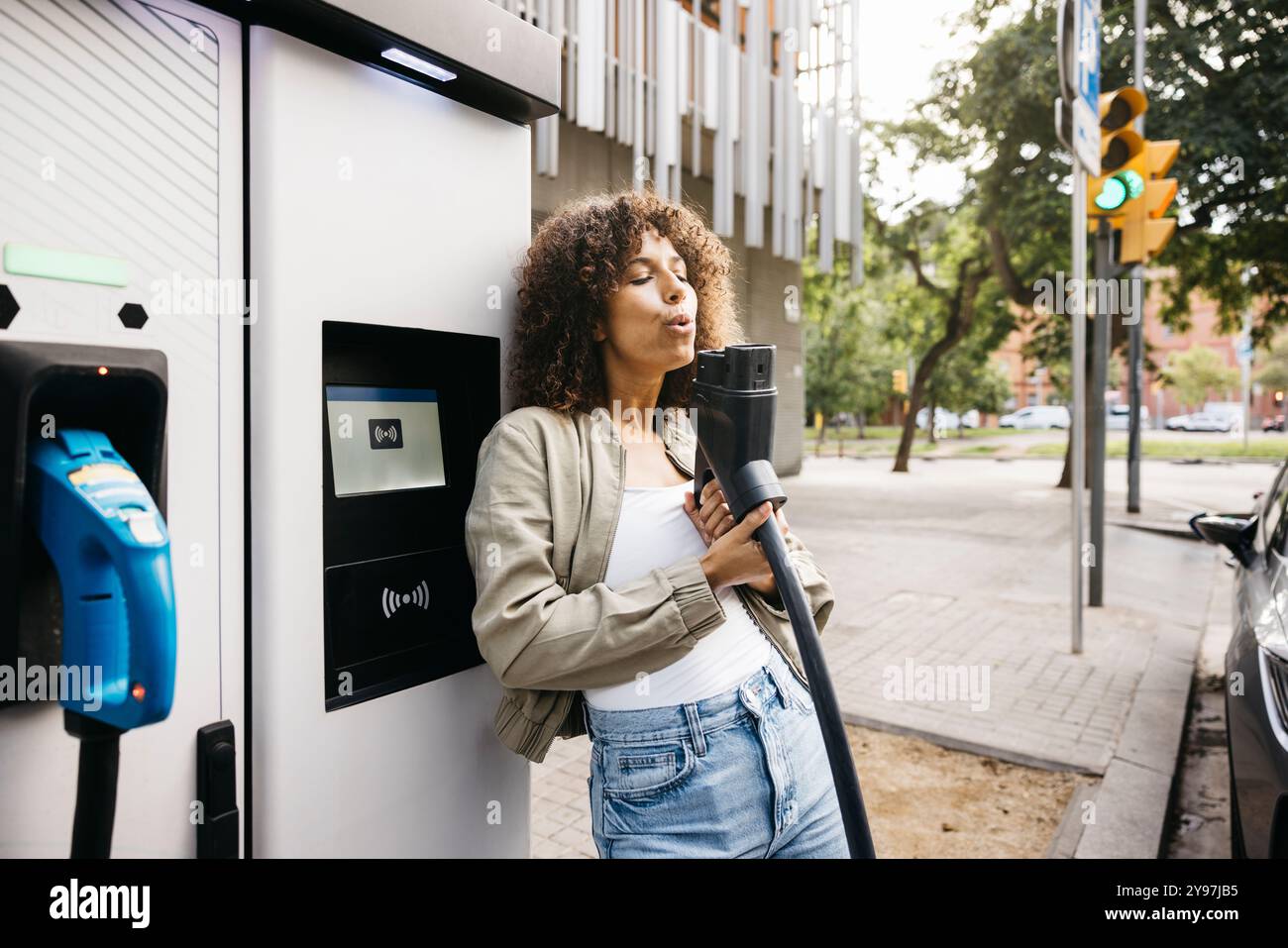 Fröhliche Frau, die ein Ladekabel als Waffe an einer Ladestation benutzt, während sie ihr Elektroauto auflädt. Frau, die umweltfreundliche Transporte und so umgeht Stockfoto