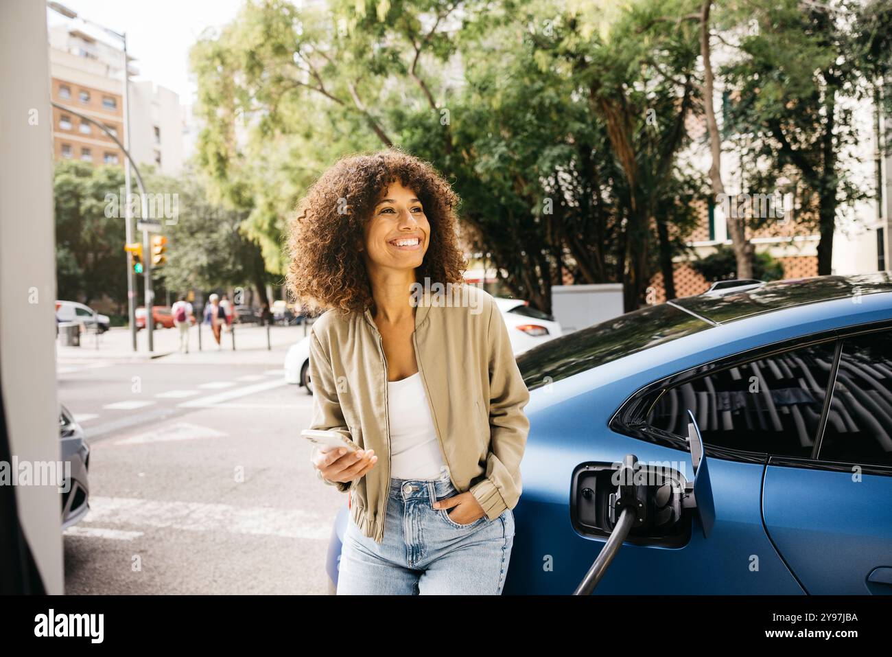 Eine Frau lächelt, als sie neben ihrem Elektroauto an einer Ladestation in einer städtischen Umgebung steht. Die Szene zeigt moderne Verkehrsmittel und ec Stockfoto