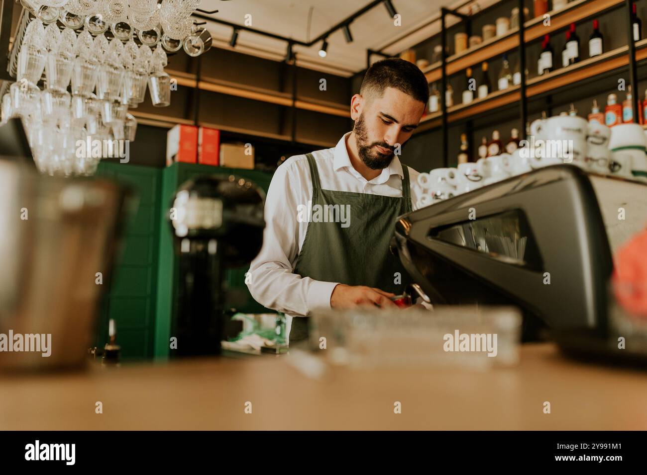 In einem schicken Café konzentriert sich ein erfahrener Barista auf die Herstellung köstlicher Getränke, umgeben von ordentlich angeordneten Gläsern und lebendiger Einrichtung, die eine warme Atmosphäre schaffen Stockfoto