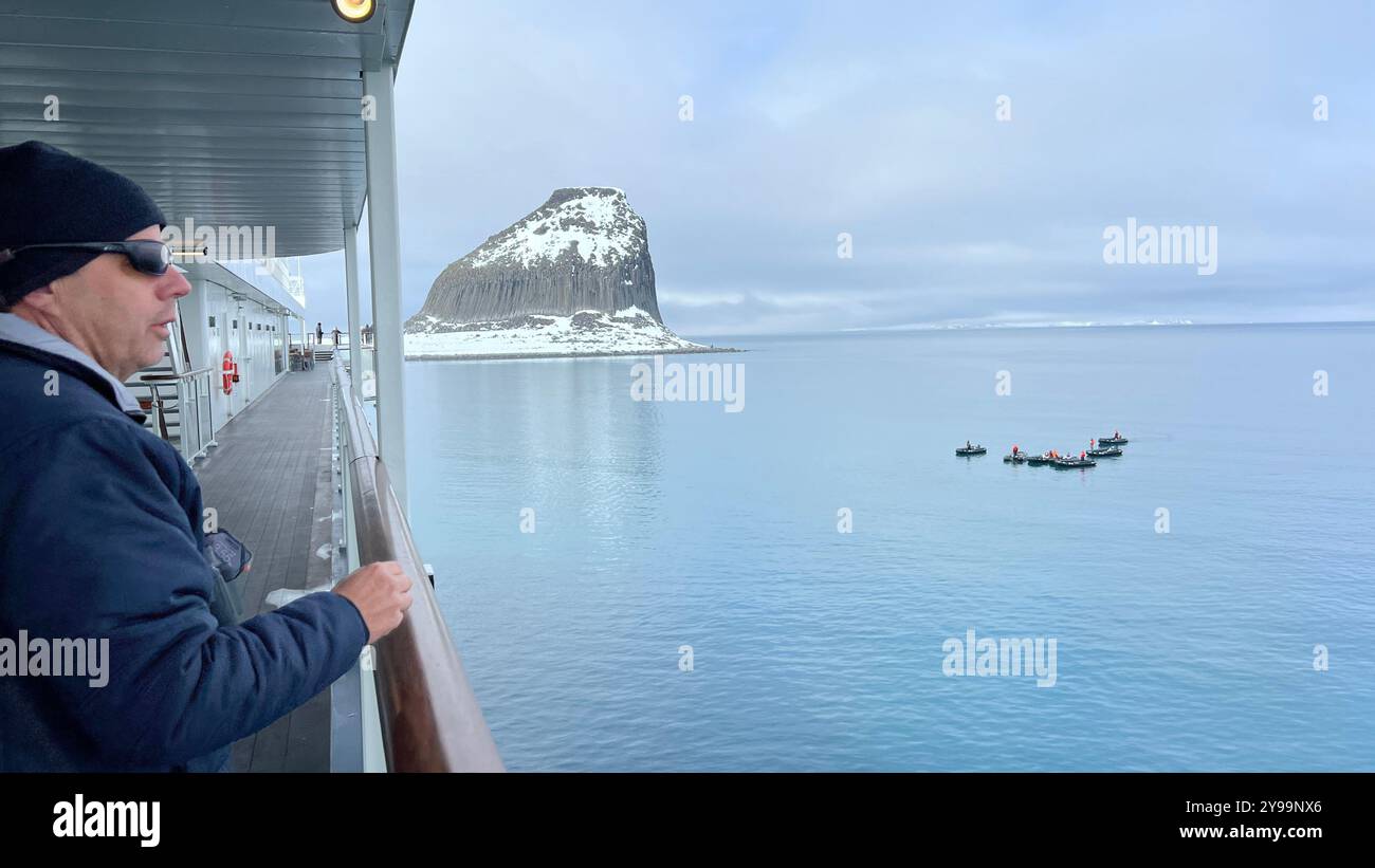 Ein Passagier auf der Lindblad Nat Geo Endurance genießt den Blick auf den Edinburgh Rock in der Antarktis, während kleine Expeditionsboote erkunden - Smartphone-aufgenommenes Stockfoto
