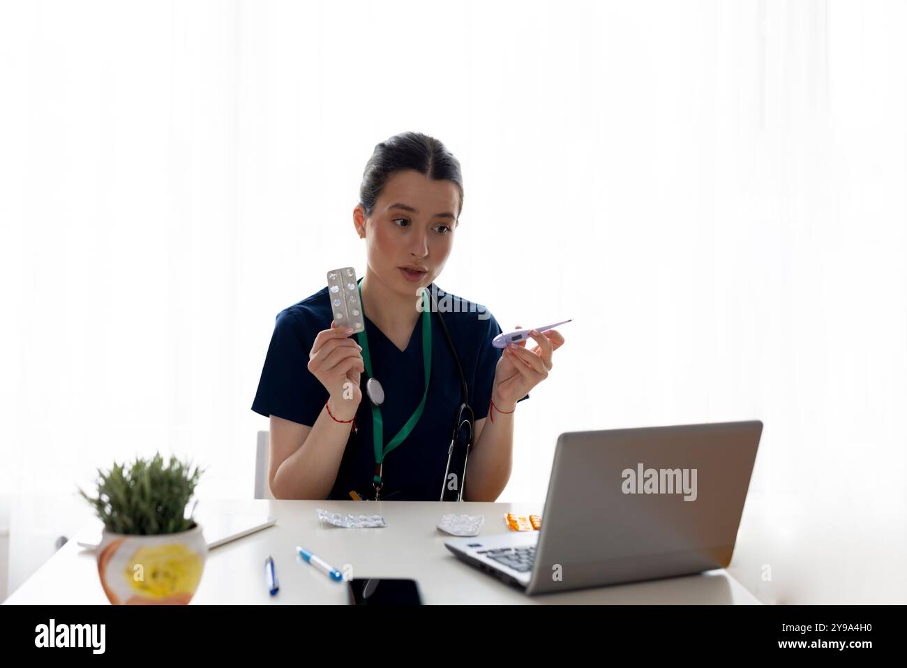 Eine junge Ärztin, die an einem Laptop über Videoanruf an einem Schreibtisch arbeitet und Pillen und Thermometer hält, Gesundheitskonzept Stockfoto