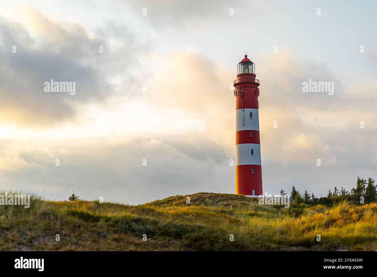 Der leuchtende Leuchtturm, in Rot und weiß gestrichen, überblickt eine ruhige Landschaft bei Sonnenuntergang, wirft warme Töne über den Himmel und beleuchtet den Himmel Stockfoto