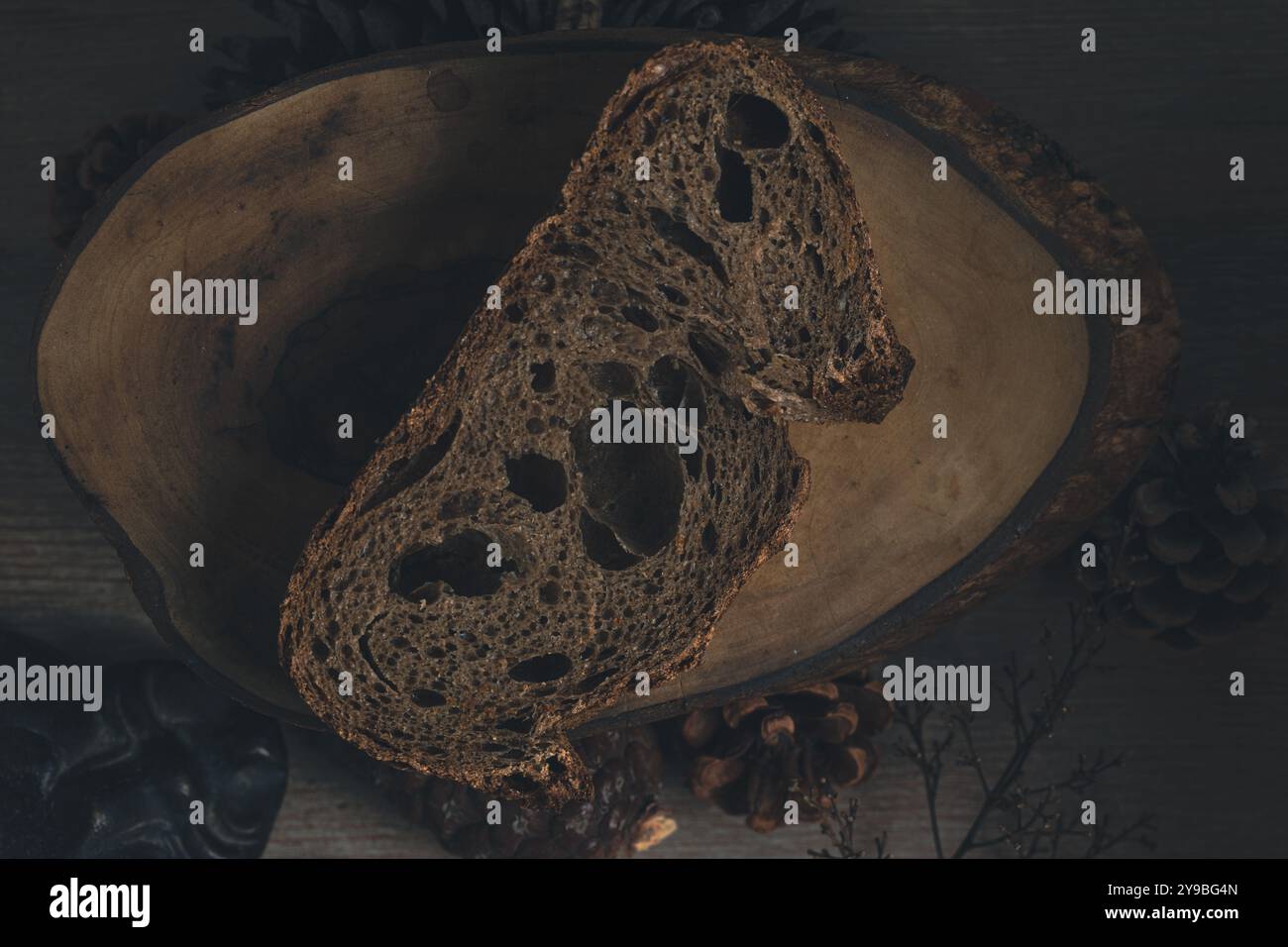 Dunkle und stimmungsvolle Aufnahme einer Scheibe Buchweizenbrot auf Holztisch mit Kegeln und trockenen Ästen, die die Herbstsaison betonen Stockfoto