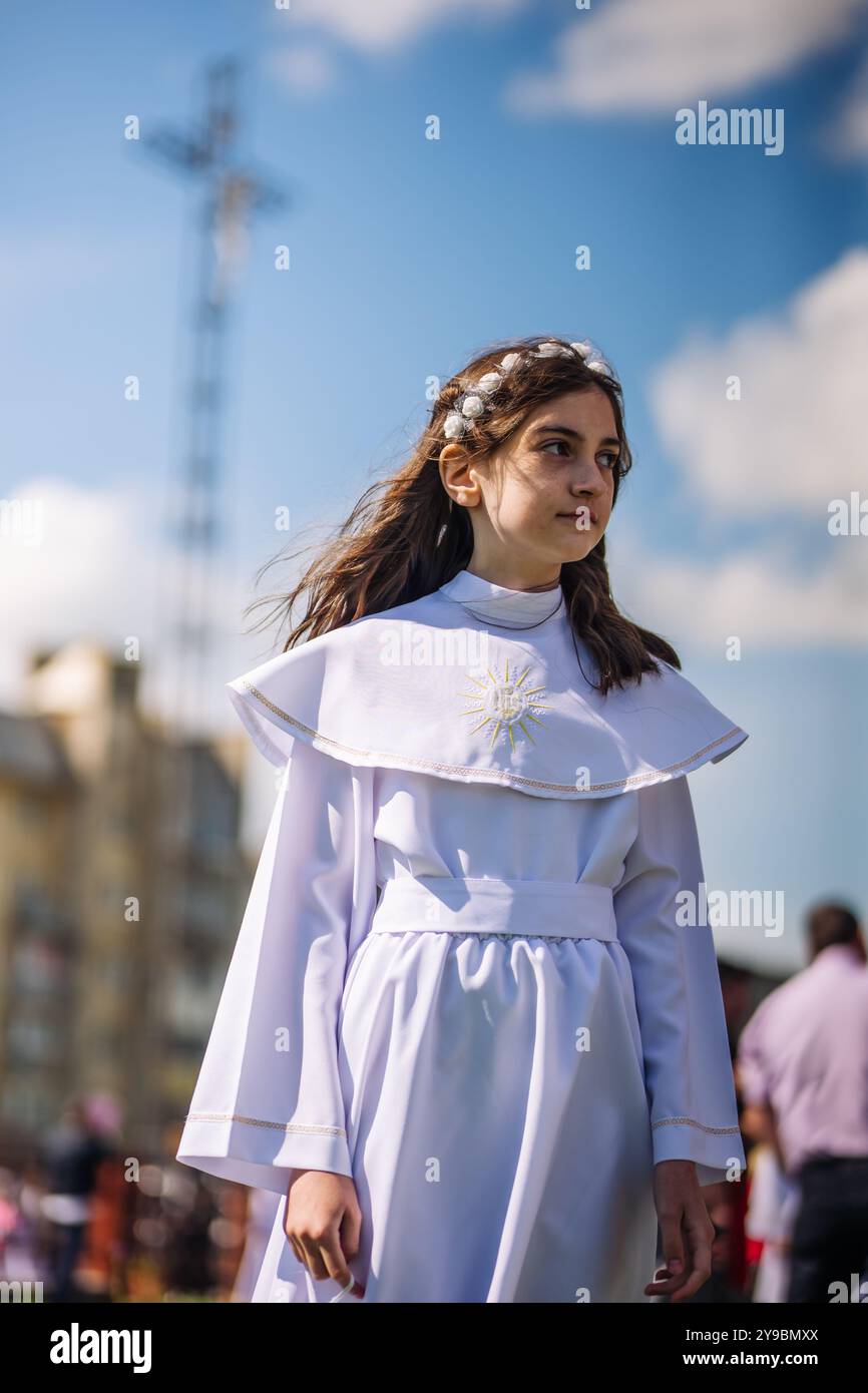 Lida, Weißrussland - 15. Juni 2021: Mädchen mit weißem Kleid und Blumenkrone im Freien mit Kreuz im Hintergrund Stockfoto