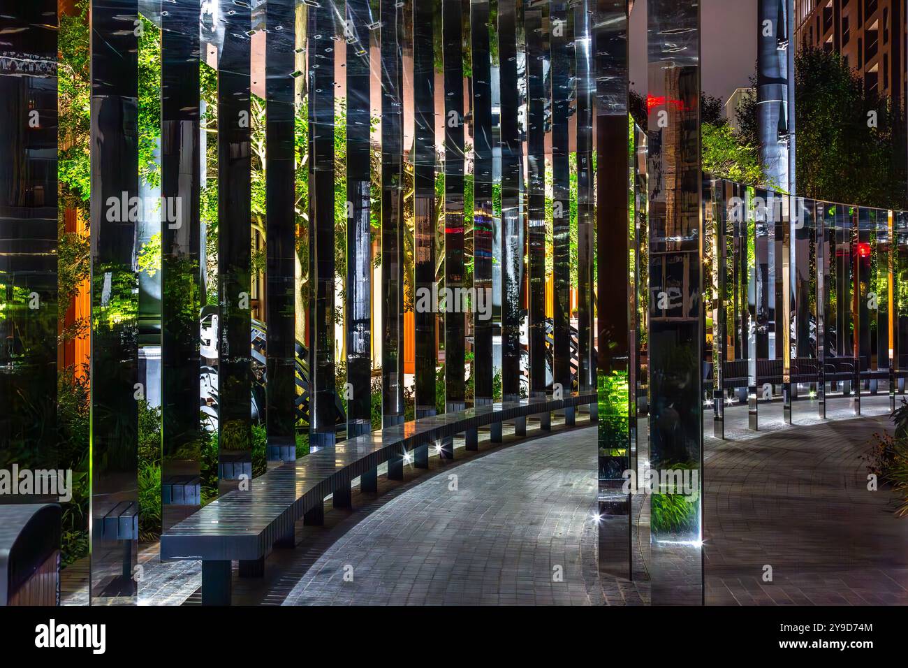 Ein moderner, architektonischer Weg, umgeben von Grün bei Nacht mit beleuchteten Bänken und reflektierenden Oberflächen in einem gut beleuchteten Stadtgebiet Stockfoto Ein moderner, architektonischer Weg, umgeben von Grün bei Nacht mit beleuchteten Bänken und reflektierenden Oberflächen in einem gut beleuchteten Stadtgebiet Stockfoto