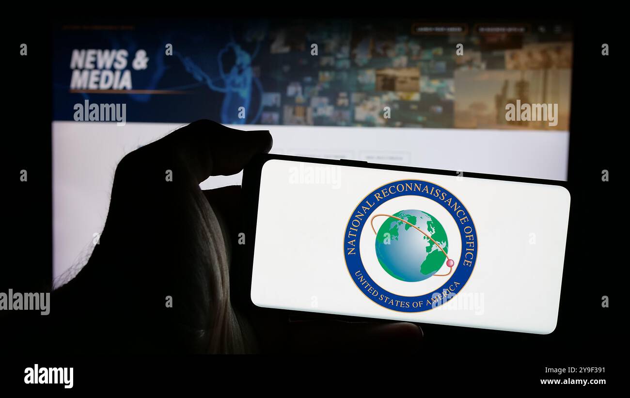 Person, die ein Mobiltelefon mit dem Logo des National Reconnaissance Office (NRO) der US-Agentur auf dem Bildschirm vor der Webseite hält. Schwerpunkt auf dem Display des Telefons. Stockfoto