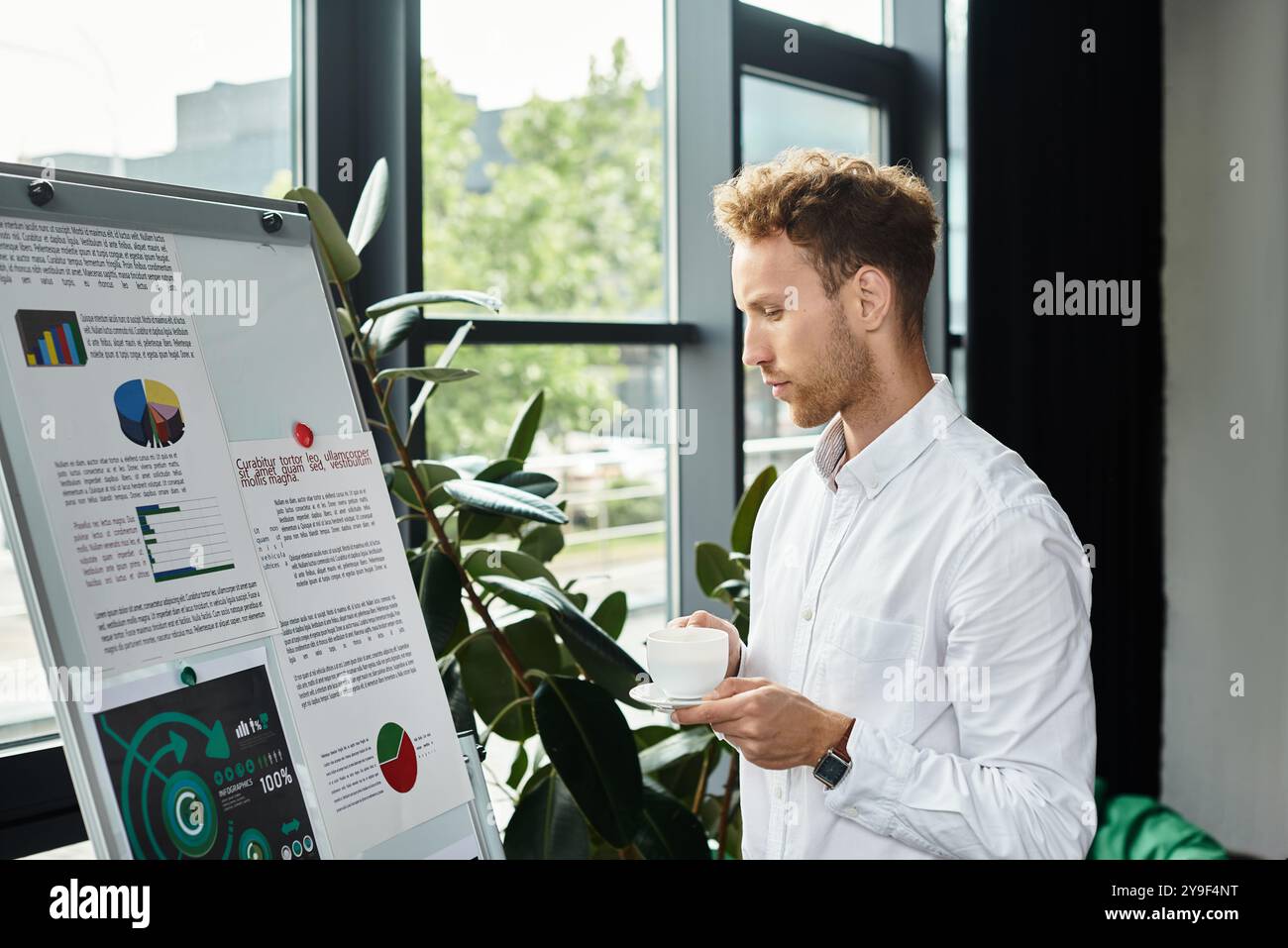 Eine dedizierte Rothaarige in einem weißen Hemd analysiert visuelle Daten, während sie in einem eleganten Büro eine Tasse Kaffee genießt. Stockfoto