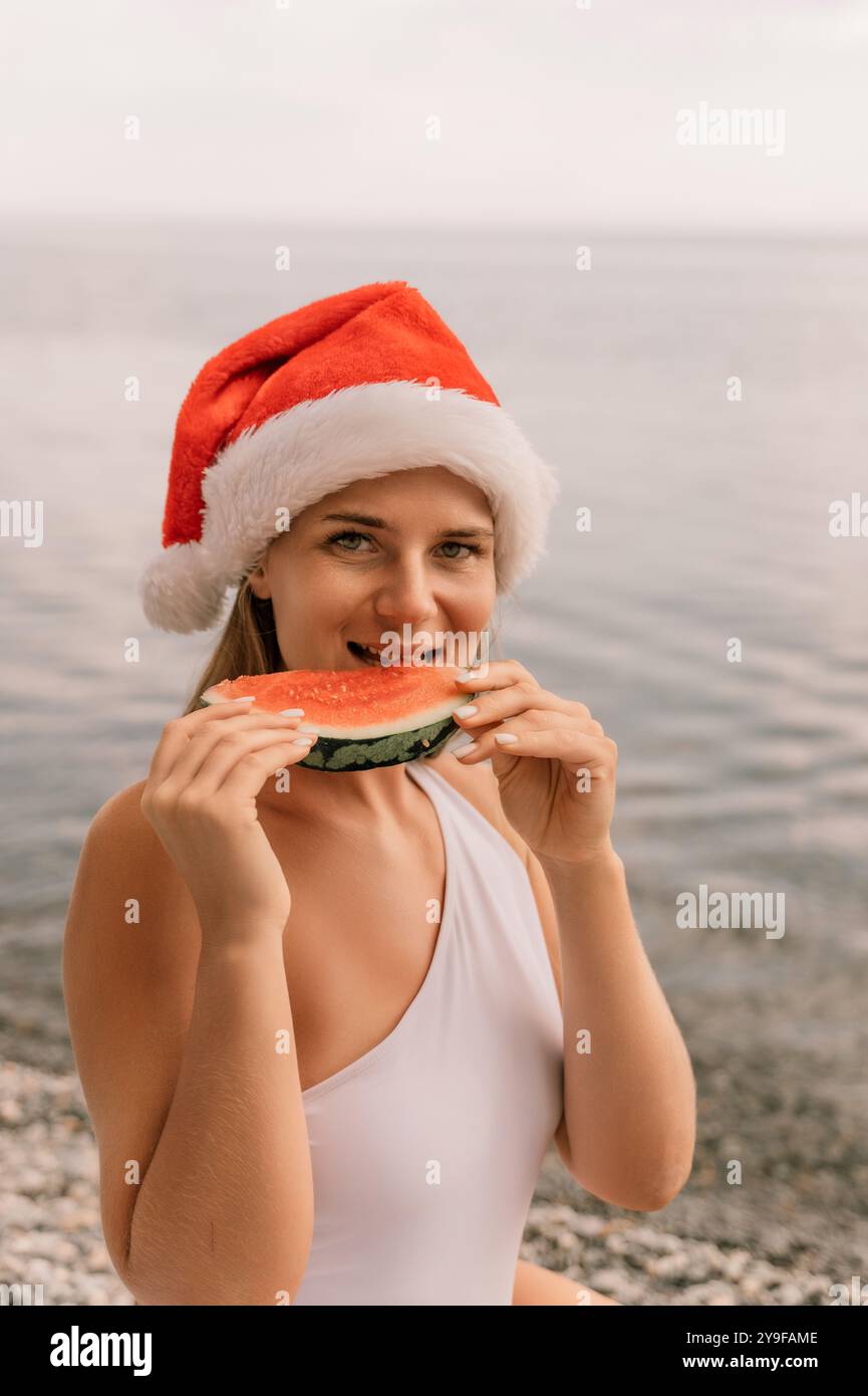 Eine Frau isst eine Wassermelone, während sie einen roten weihnachtsmann-Hut trägt. Die Szene ist unbeschwert und festlich, während die Frau einen erfrischenden Snack auf einem genießt Stockfoto