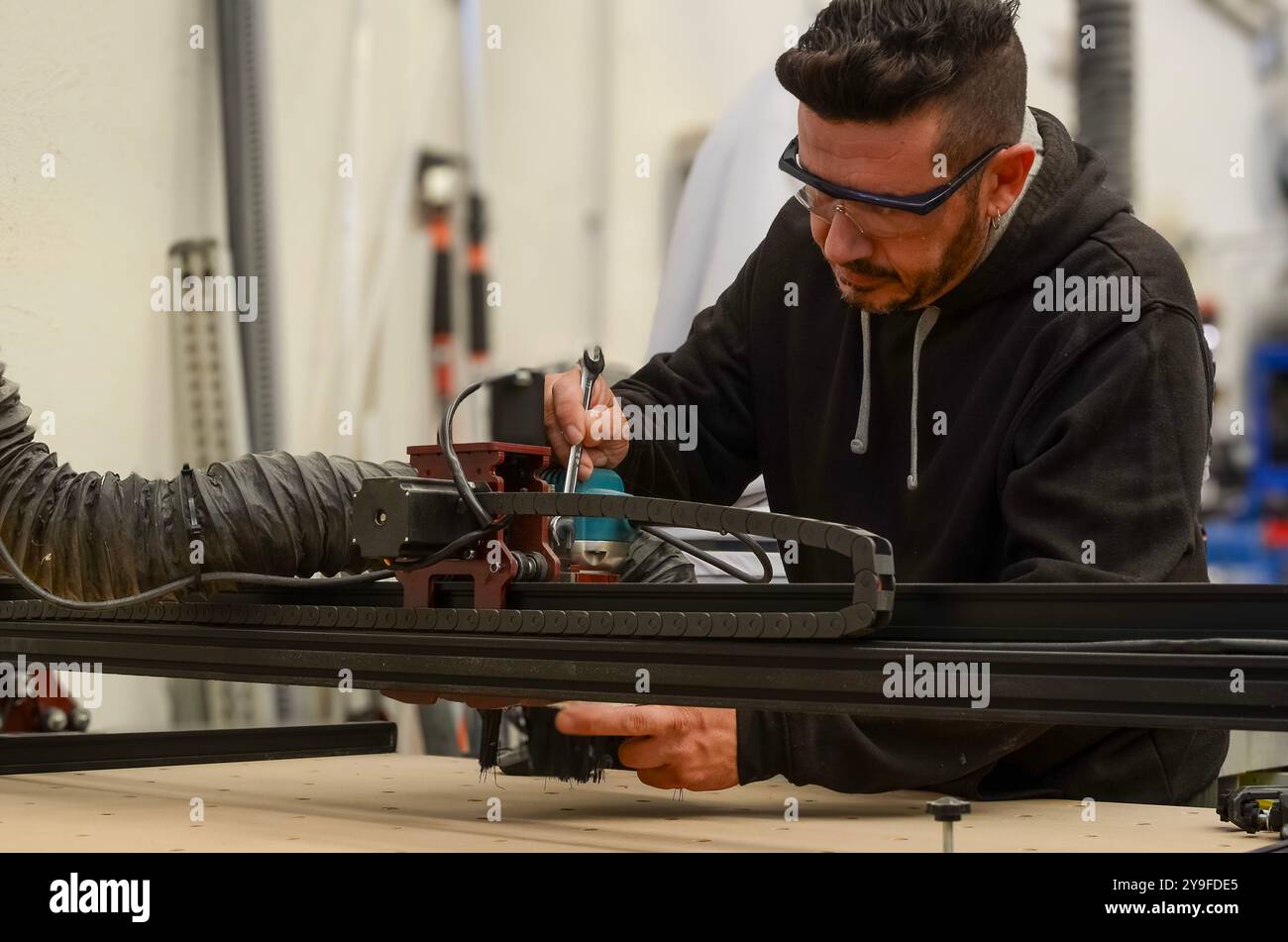 Wartungstechniker reparieren eine numerische Steuerung zum Schneiden von Holz mit einem Schraubenschlüssel in der Hand, einer Schutzbrille und einem schwarzen Sweatshirt Stockfoto