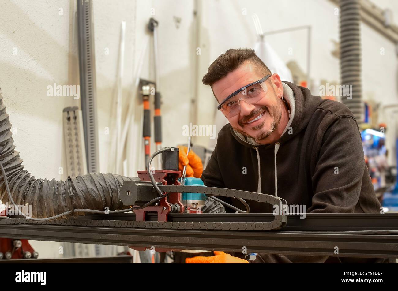 Instandhaltungstechniker reparieren eine numerische Steuerung zum Schneiden von Holz mit einem Schraubenschlüssel in der Hand, einer Schutzbrille, einem schwarzen Sweatshirt und einem orangefarbenen Handschuh Stockfoto