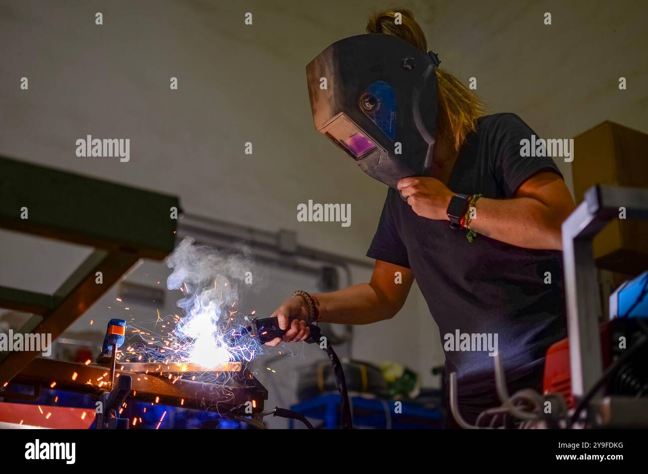 Frau schweißt Eisen mit Elektroschweißgerät in der Werkstatt, tragend Schutzausrüstung Stockfoto