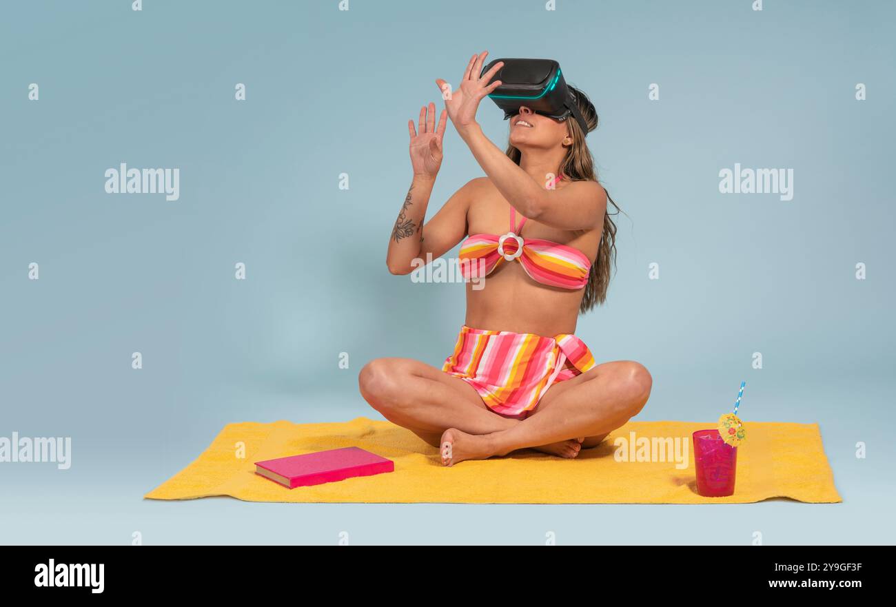 Porträt der schönen brünetten Latina Frau mit langen Haaren, die eine Virtual-Reality-Brille trägt, auf einem gelben Handtuch sitzt, rot-gelb gestreifte BI trägt Stockfoto