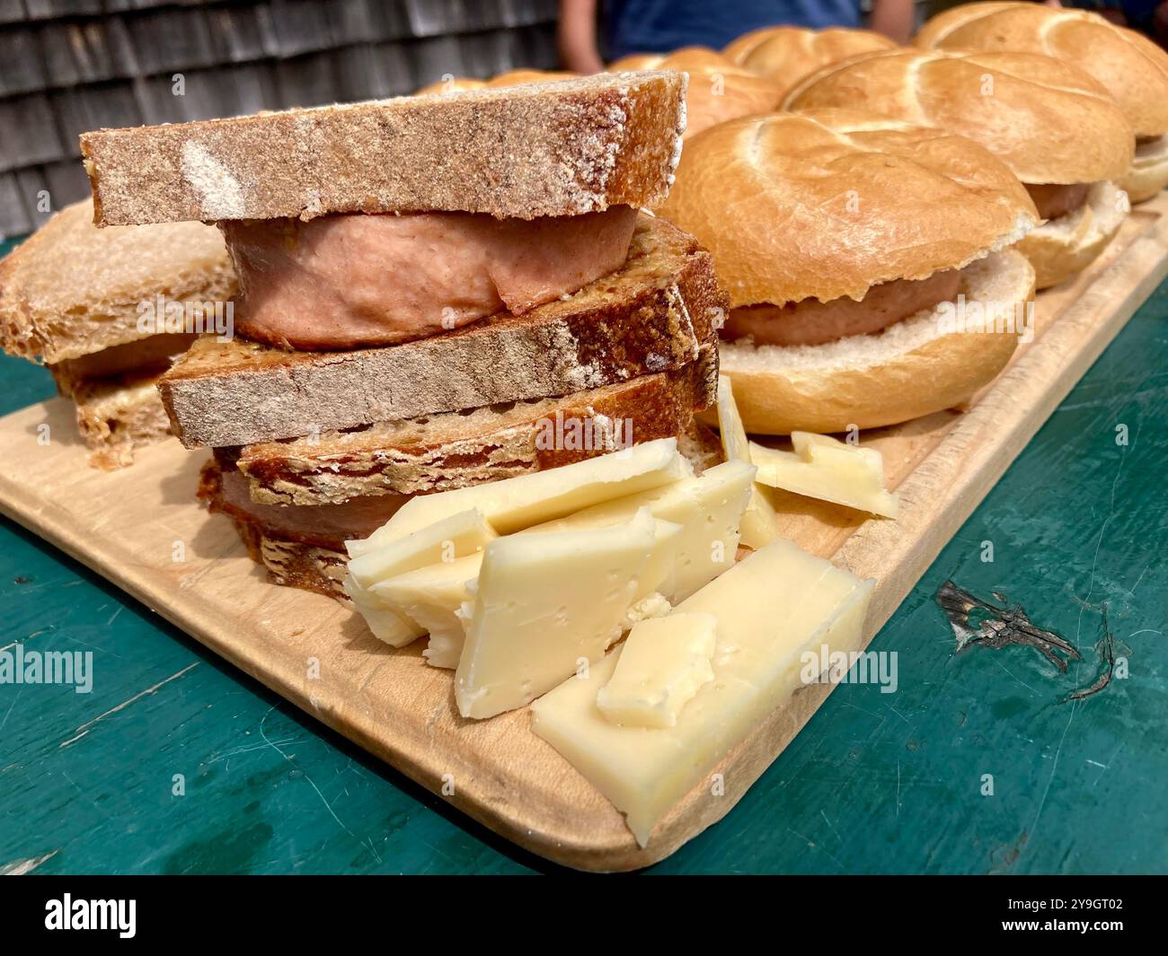 Ein hölzernes Schneidebrett auf einem grünen Tisch mit Leberkaese-Brötchen, Brot und Käsestücken Stockfoto