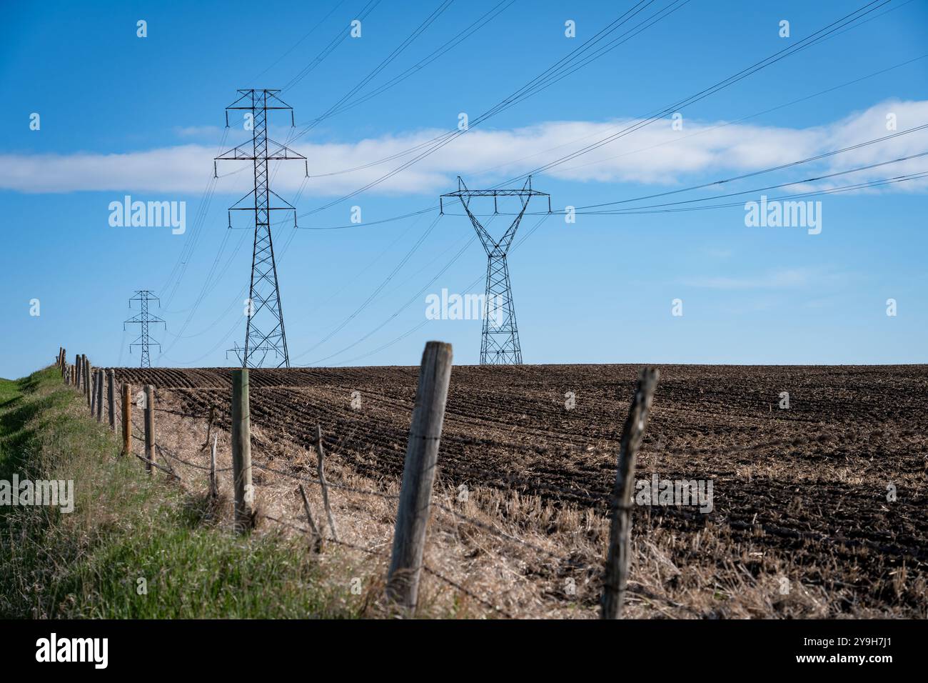 Stromleitungen, die von hohen Stahlgittertürmen auf einem gepflügten Feld mit Stacheldrahtzaun auf den kanadischen Prärien in Rocky View Count hoch gehalten werden Stockfoto