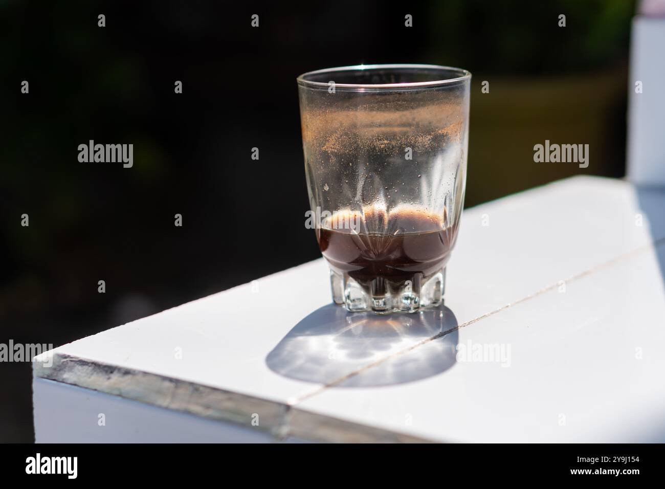 Ein Glas mit dunkler Flüssigkeit, möglicherweise Kaffee, steht auf einer weißen Oberfläche. Das Glas ist teilweise gefüllt und zeigt Reste des Getränks. Der Hintergrund Stockfoto