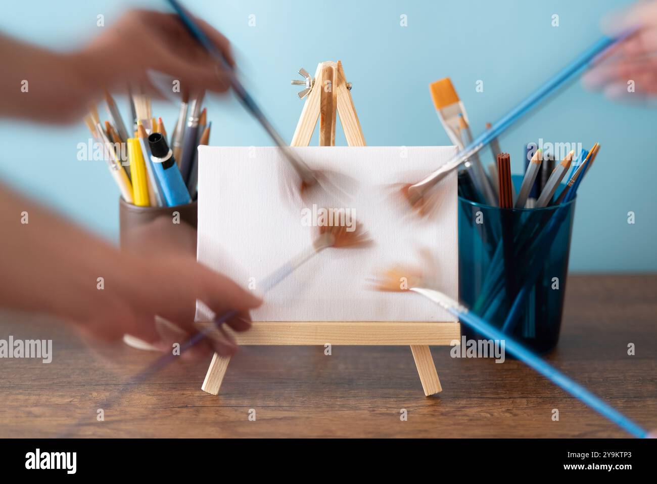 Vorderansicht des sich bewegenden Pinsels, der Pinselstriche auf kleiner Holzstaffelei mit leerer Leinwand auf Holztisch mit Glas mit Farbtuben macht, br Stockfoto