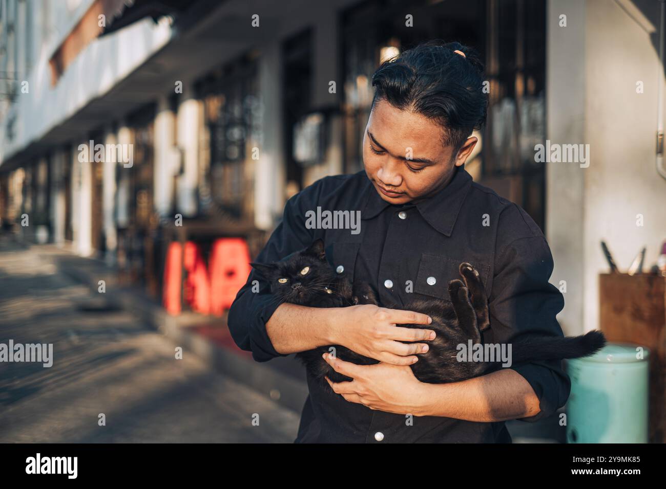 Ein südostasiatischer Mann, der eine schwarze Katze mit Bokeh-Hintergrund wiegt Stockfoto