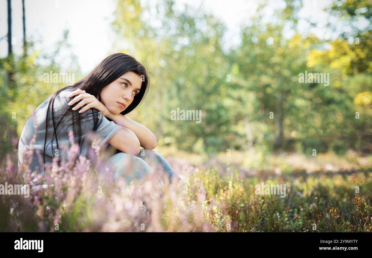 Eine junge Frau mit langen dunklen Haaren sitzt nachdenklich zwischen blühenden Wildblumen, umgeben von einem üppigen grünen Wald, und genießt einen friedlichen Nachmittag im Freien Stockfoto