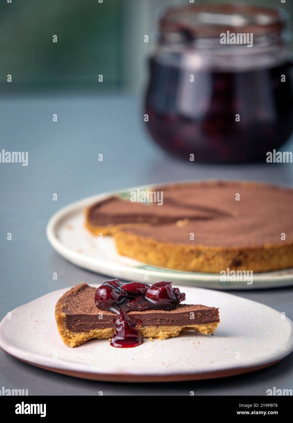 Ein Stück Schokoladenkuchen mit ganzer Torte und einem Glas Kirschen Stockfoto