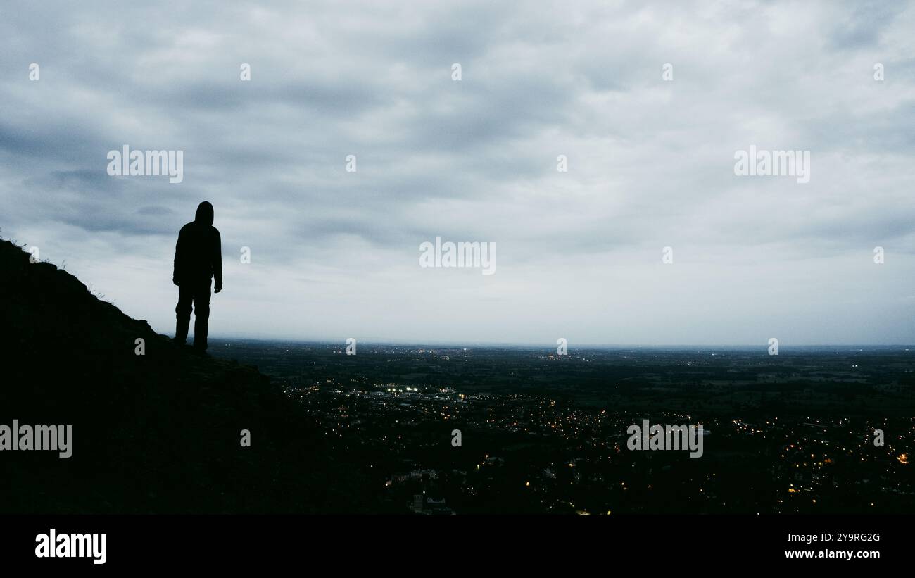 Eine geheimnisvolle Figur mit Kapuze, die auf einem Hügel steht und auf eine Stadt blickt, deren Straßenlaternen am Abend leuchten. Stockfoto