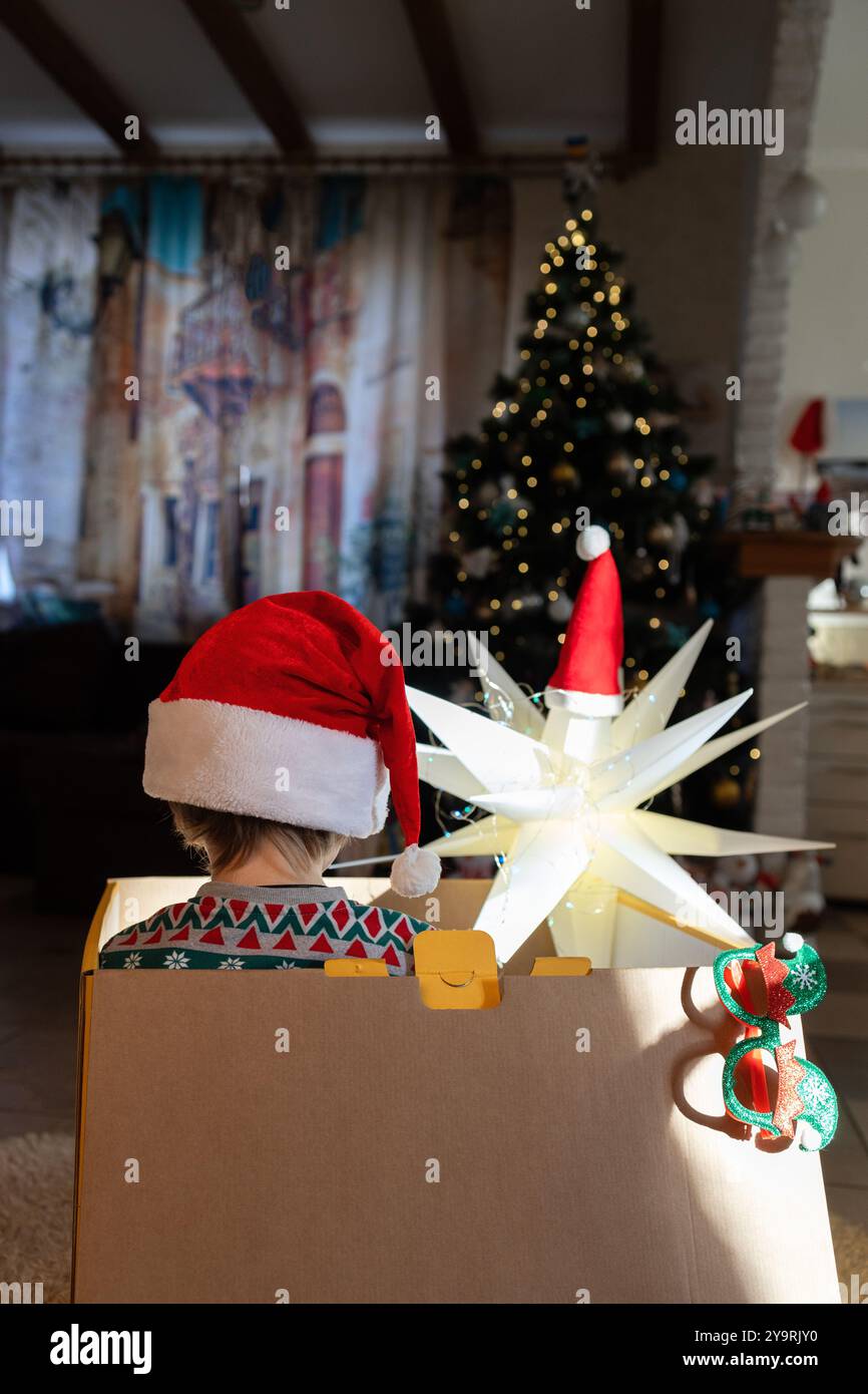 Unerkennbares Kind im Weihnachtsmann-Hut sitzt in einem großen Karton mit leuchtender Laterne im Zimmer vor dem Weihnachtsbaum. Warten auf das Weihnachtswunder. Stockfoto