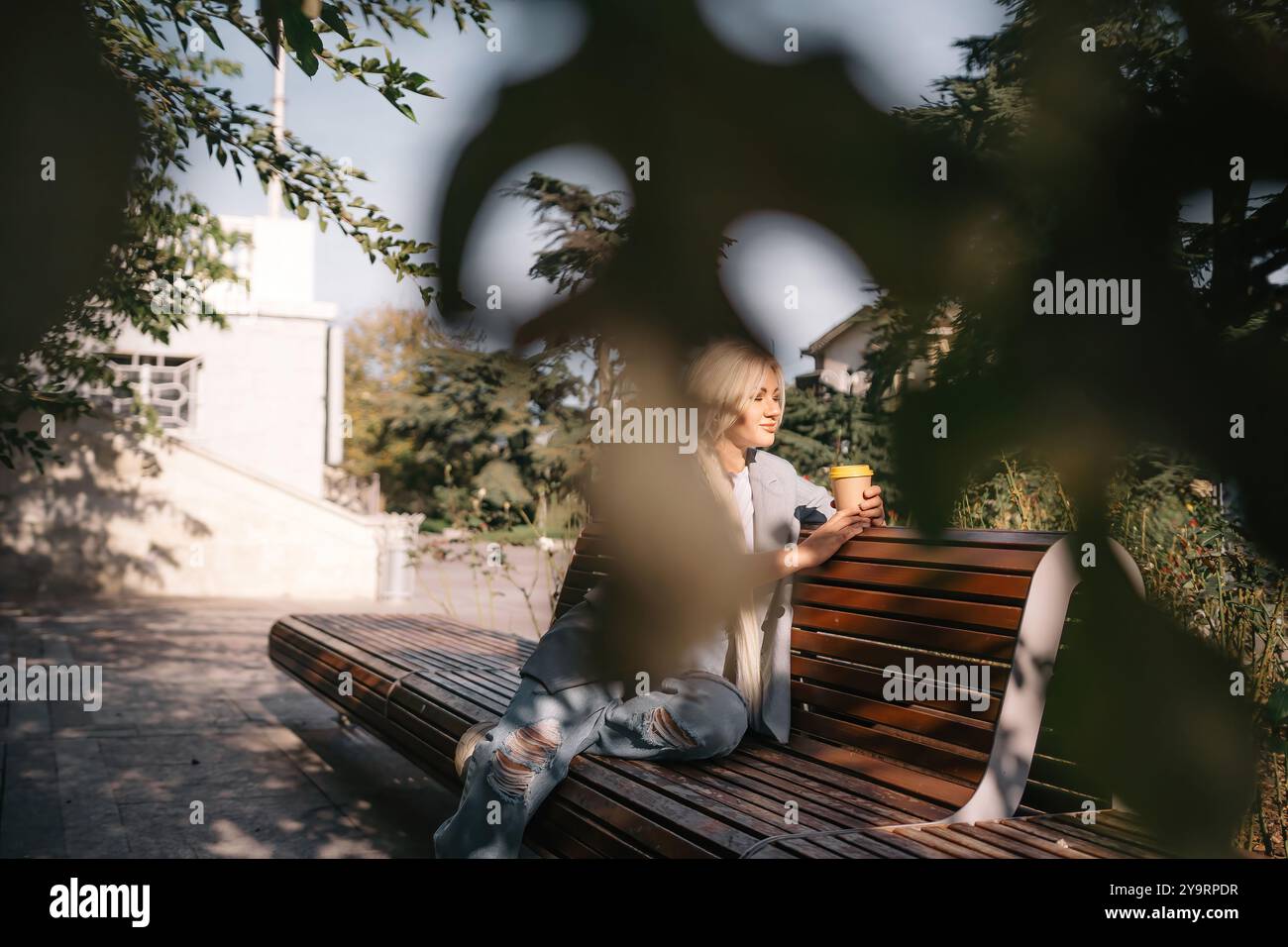 Eine blonde Frau sitzt auf einer Bank mit einer Tasse Kaffee in der Hand. Sie trägt eine graue Jacke und Jeans. Die Bank ist aus Holz und hat einen geschwungenen Rücken Stockfoto