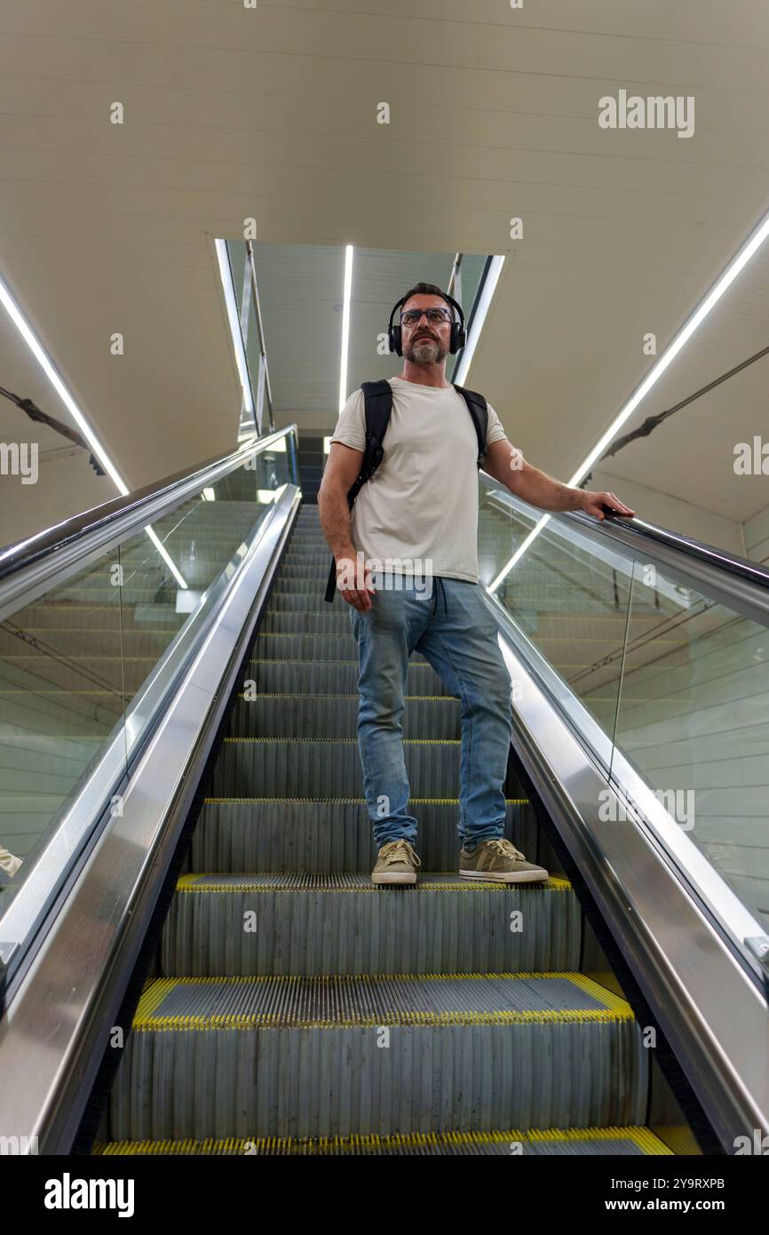 Mann mittleren Alters in lässiger Kleidung mit Rucksack, der am Bahnhof auf dem Eskalator absteigt, um die Bahnsteige zu erreichen Stockfoto