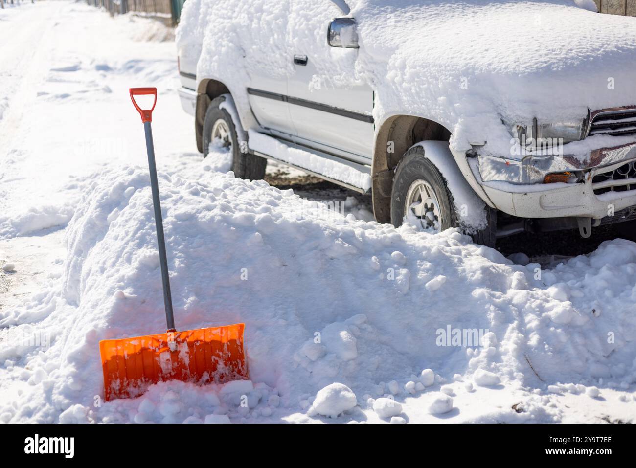 Die orangefarbene Schneeschaufel ragt aus der Schneewolke vor dem schneebedeckten weißen SUV-Auto hervor Stockfoto