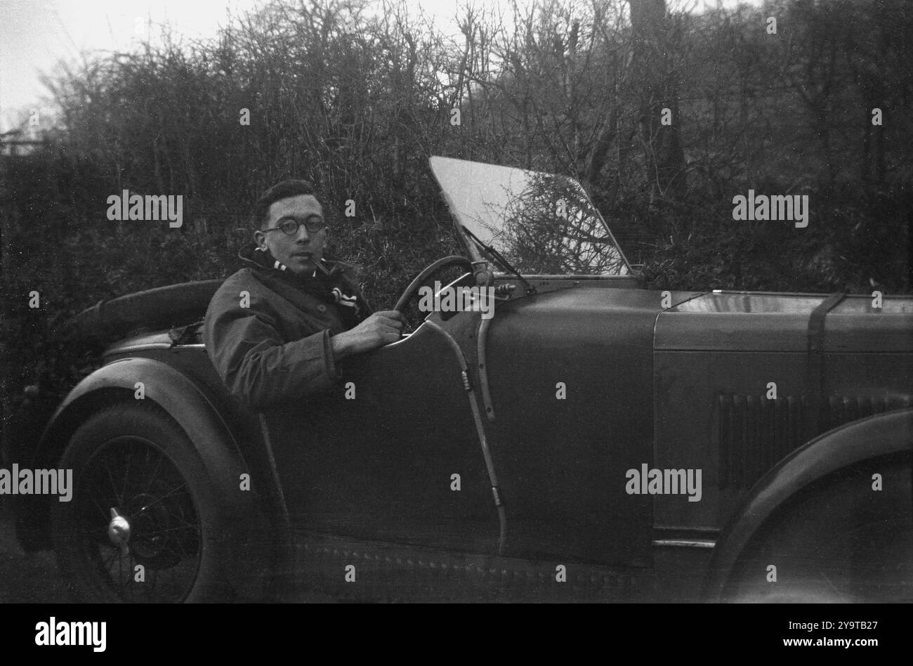 1950er Jahre, historisch, ein Gentleman, Pfeife im Mund, Schal um den Hals, Mantel an, sitzt in seinem offenen Sportwagen der Epoche. Der Zweisitzer verfügt über ein Reserverad auf der Rückseite, ein Lederband um die Motorhaube und Speichenräder. Stockfoto