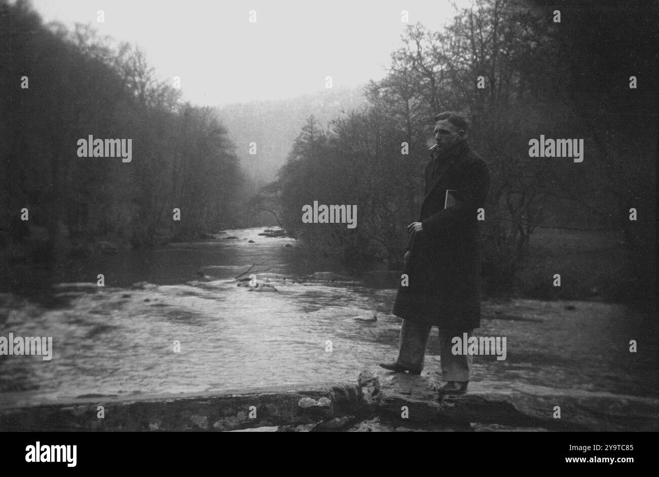 1960er Jahre, historisch, draußen in einem Fluss in einem Wald, ein Mann in einem Mantel, der mitten im Wasser auf einem Felsübergang steht und raucht. Stockfoto
