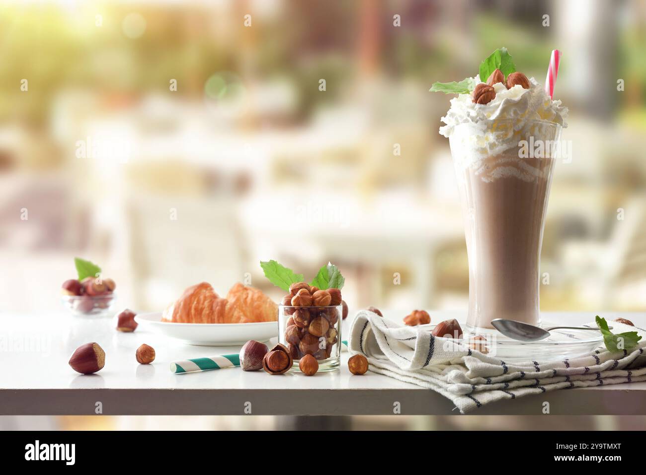 Milchshake mit Haselnüssen und Sahne, dekoriert in einem Glas auf einem Terrassentisch mit Croissant und Nüssen. Vorderansicht. Stockfoto