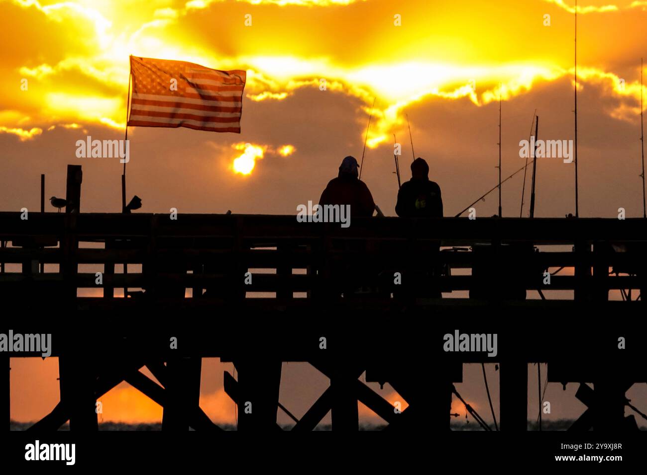 Isle Of Palms, Usa. Oktober 2024. Fischer, die unter amerikanischer Flagge stehen, umrahmt von einem dramatischen Sonnenaufgang am Isle of Palms Pier, 11. Oktober 2024 in Isle of Palms, South Carolina. Hurrikan Milton zog 400 Meilen weiter nach Süden, führte aber herbstliches Wetter entlang der Küste, während die Stürme aufwachen. Quelle: Richard Ellis/Richard Ellis/Alamy Live News Stockfoto