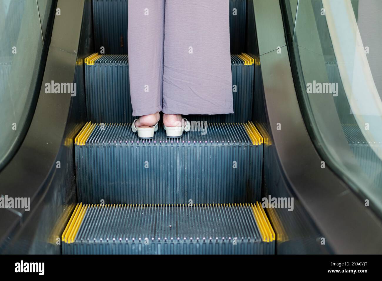 Nahaufnahme der Füße einer Person auf einer Rolltreppe. Die Person trägt helle Sandalen und lose, graue Hosen. Die Rolltreppen Stockfoto