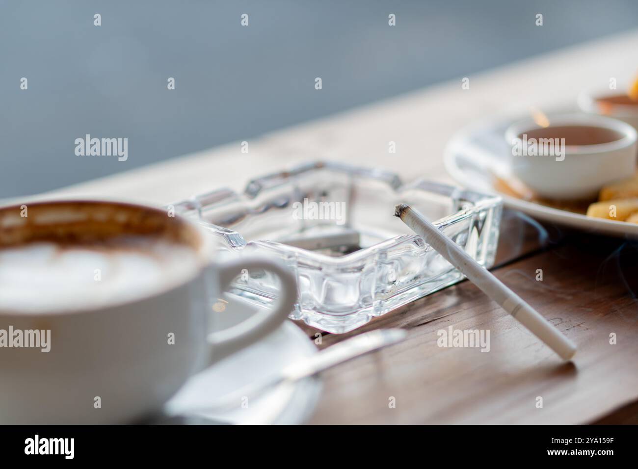 Eine gemütliche Cafeszene mit einer Tasse Kaffee, einem Aschenbecher mit einer Zigarette und einem Teller mit Snacks. Die Umgebung hat eine warme und einladende Atmosphäre, Perfe Stockfoto