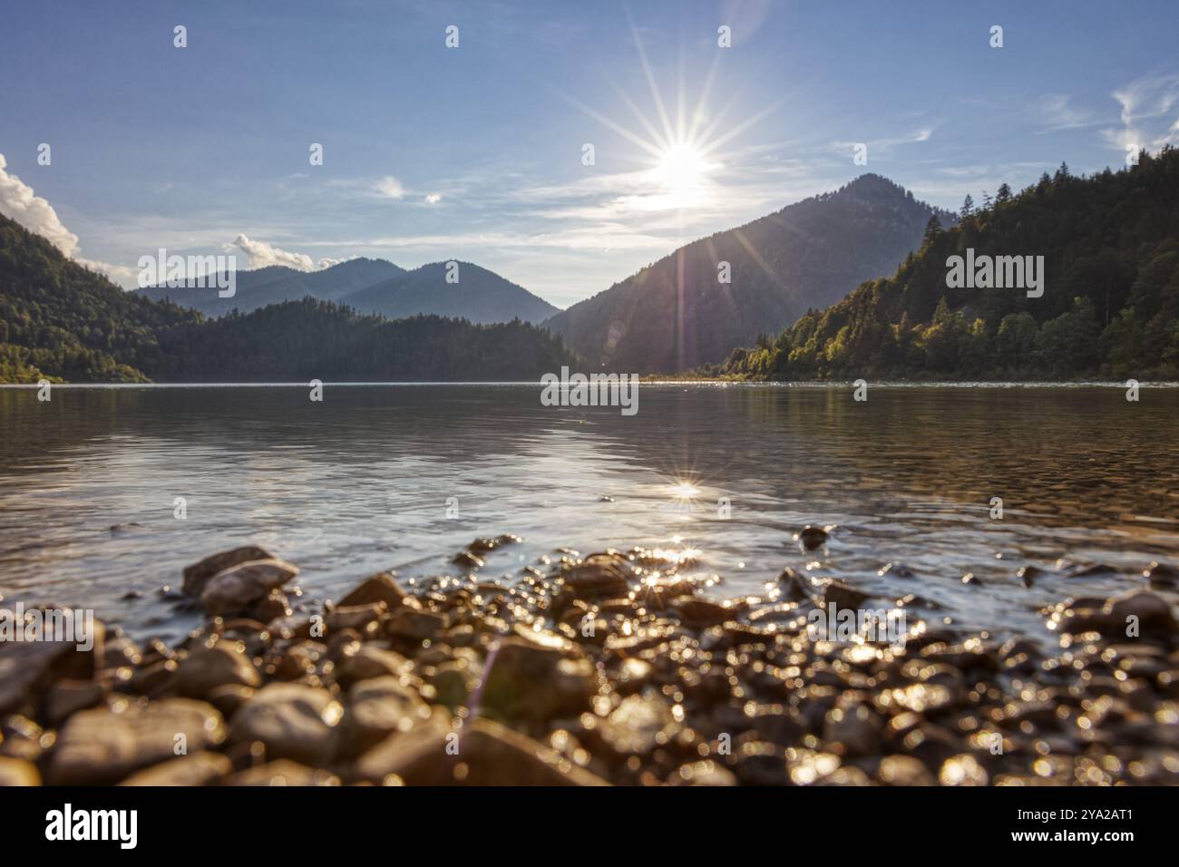 Ruhiger See mit Bergkulisse, goldenem Himmel und Sonnenlicht auf Wasser und Steinen, Weitsee Stockfoto