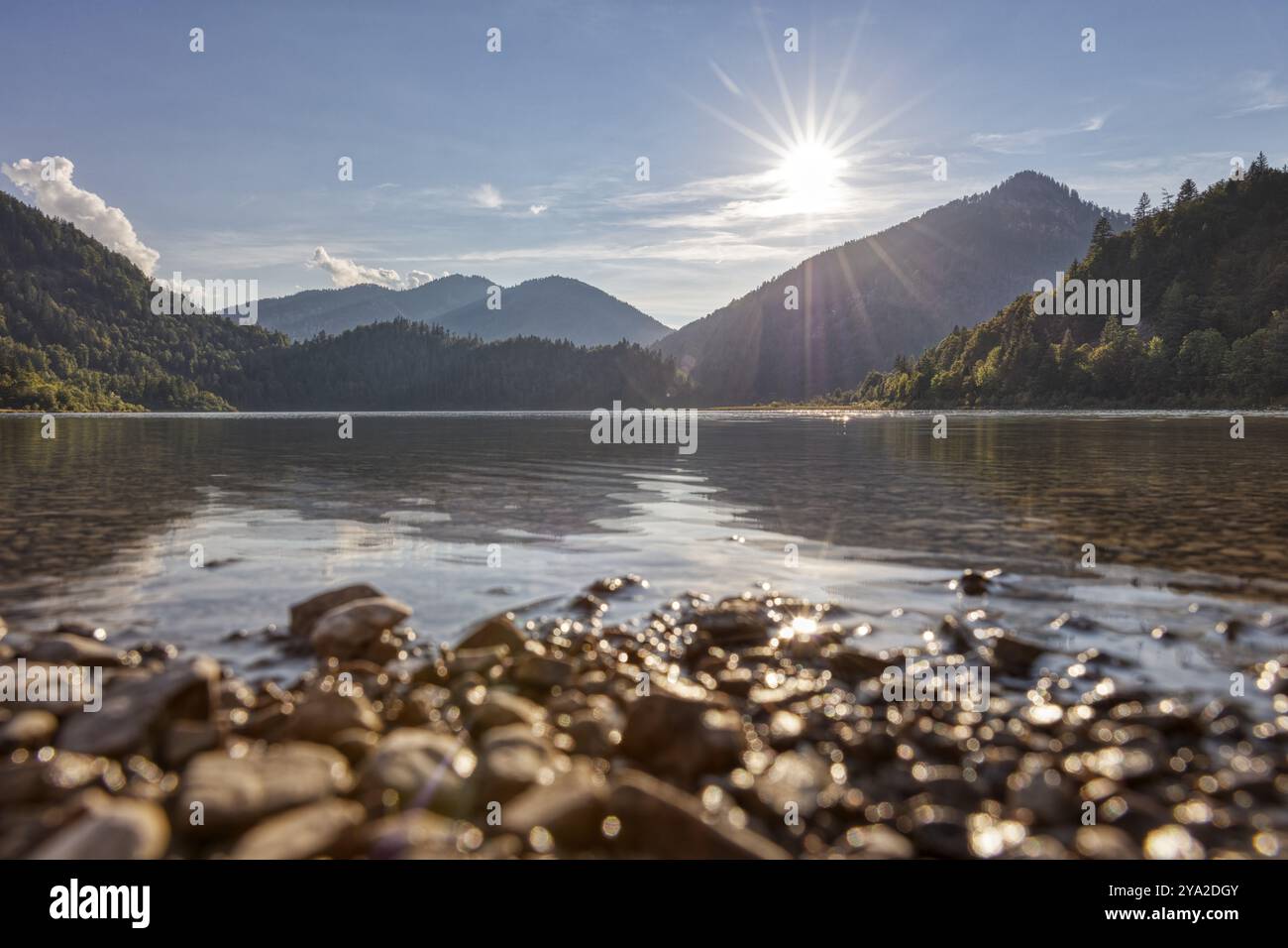 Ruhiger See mit Bergkulisse, goldenem Himmel und Sonnenlicht auf Wasser und Steinen, Weitsee Stockfoto