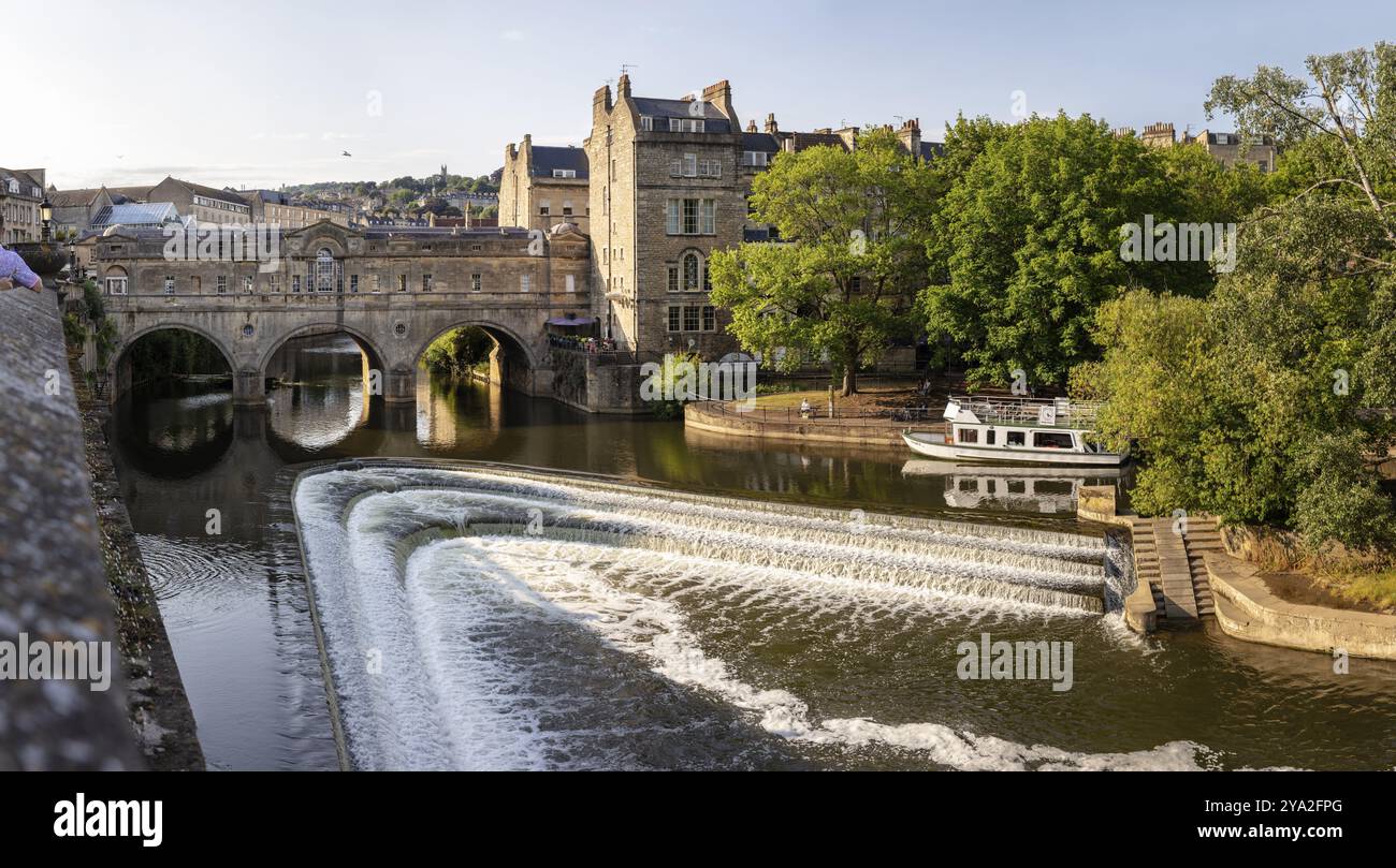Fluss mit Wasserfall und Brücke, umgeben von urbaner Landschaft und Natur, Bath Stockfoto