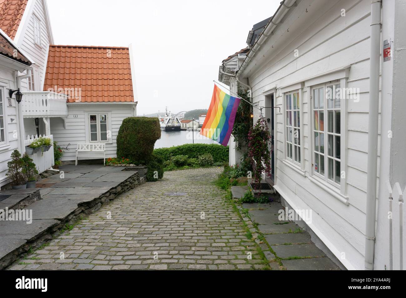 Eine ruhige Straße im historischen Viertel Gamle Stavanger, mit einem weißen Holzhaus mit einer LGBT-Regenbogenfahne, die an der Fassade und dem Meer hängt Stockfoto