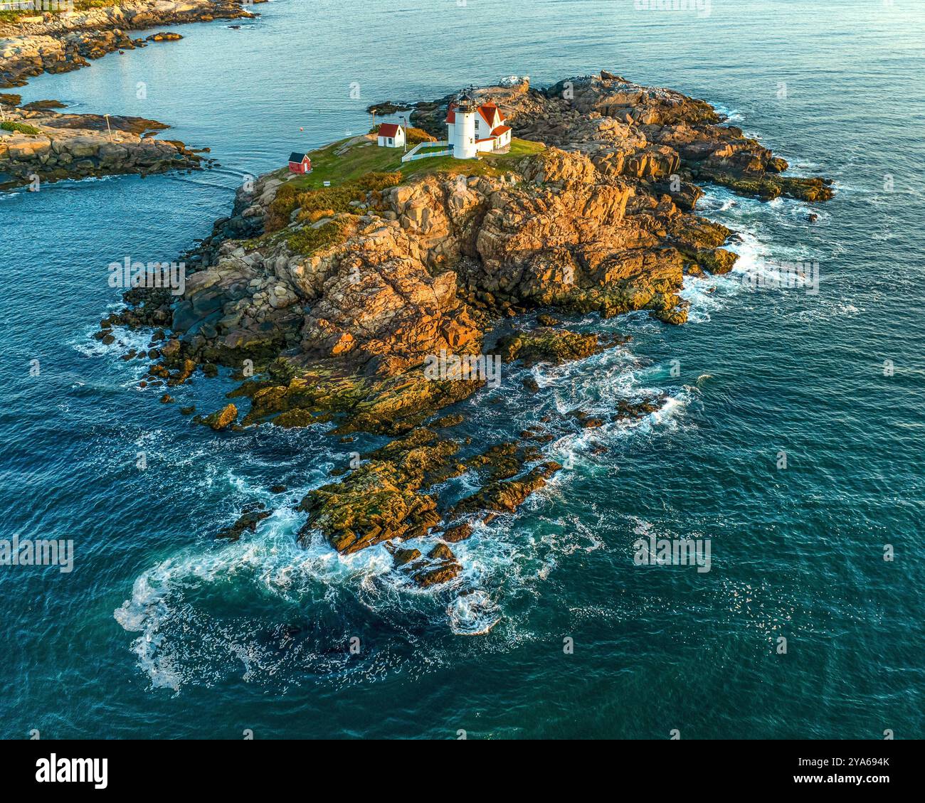 Ein Leuchtturm steht auf einer felsigen Insel, umgeben vom blauen Meer, mit Wellen, die sanft gegen die Felsen stürzen. Die Szene fängt das friedliche und ist ein Stockfoto