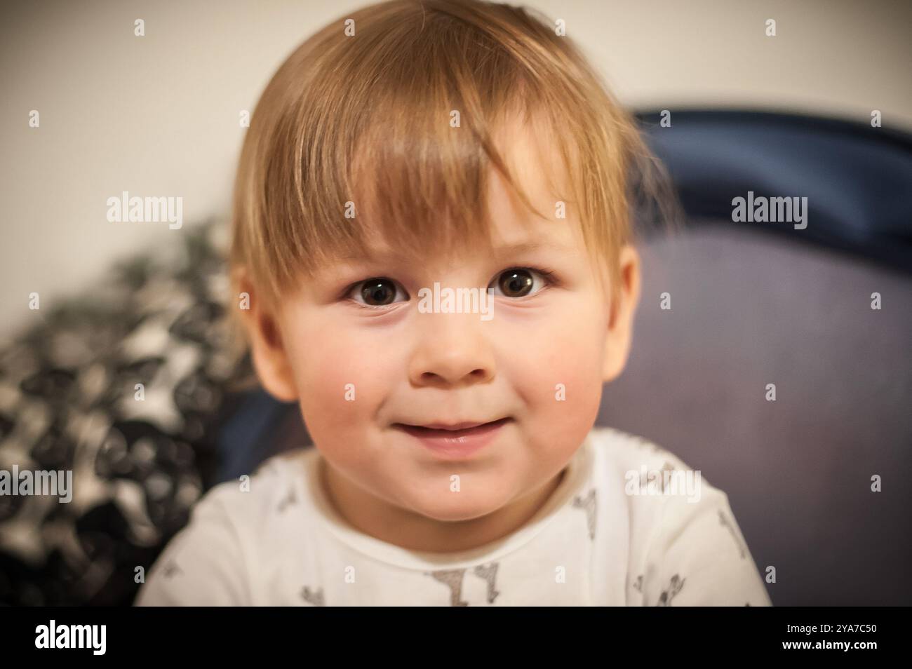 Fröhliches kleines Kind mit hellbraunem Haar blickt mit einem warmen Lächeln direkt in die Kamera. Die gemütliche Innenumgebung trägt zur fröhlichen Atmosphäre bei. Stockfoto