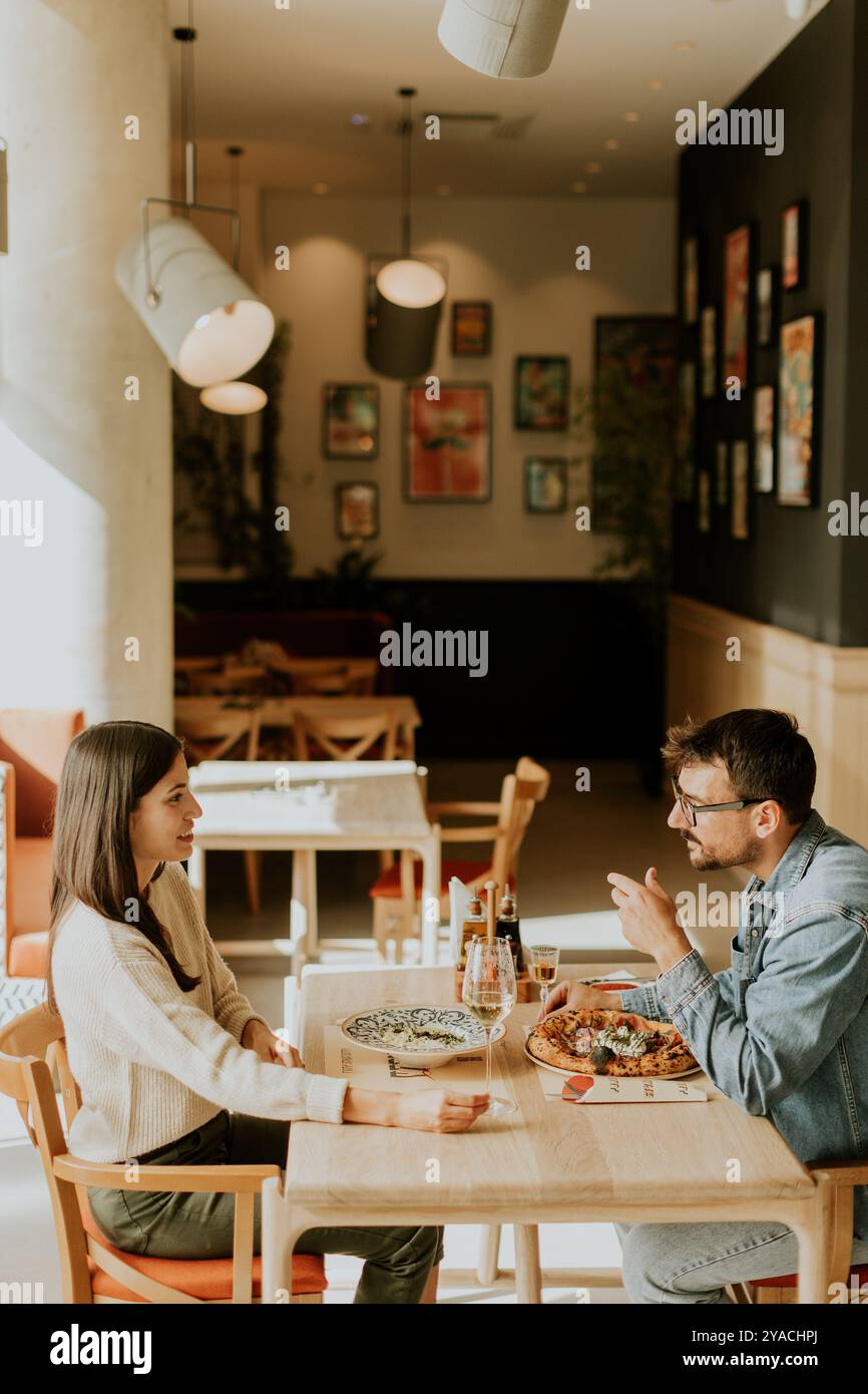 Junge Paare teilen einen herzlichen Moment in einem Café, lächeln und halten Hände über die Getränke, während sie von einer charmanten und künstlerischen Atmosphäre umgeben sind. Stockfoto