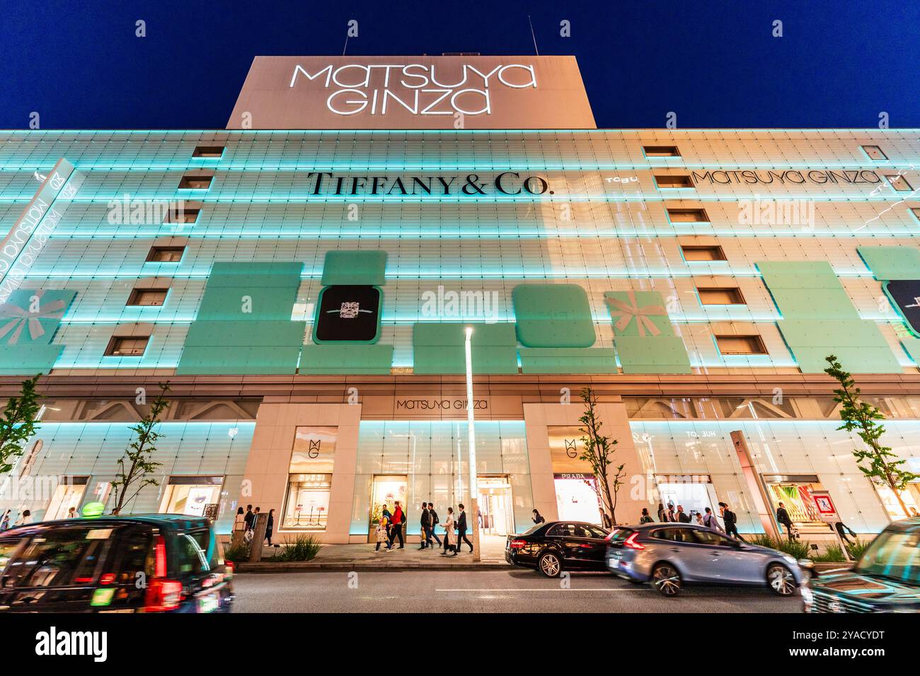 Auf der anderen Straßenseite bietet Ihnen ein flacher Blick auf das Wahrzeichen Matsuya Ginza und das Tiffany Store Building, beleuchtet in blauem, grünem Licht. Verkehr, der auf der Straße vorbeifährt. Stockfoto