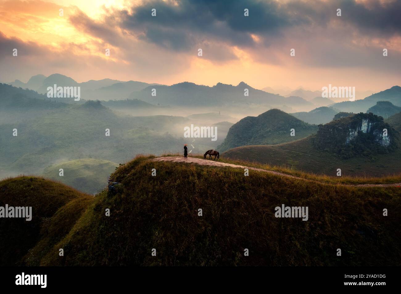 Blick aus der Vogelperspektive auf den wunderschönen Sonnenaufgang über Ba Guang Grashügel, Bergkette und Nebel zusammen mit Dorfbewohnern, die ihr Pferd auf den Berg bringen Stockfoto