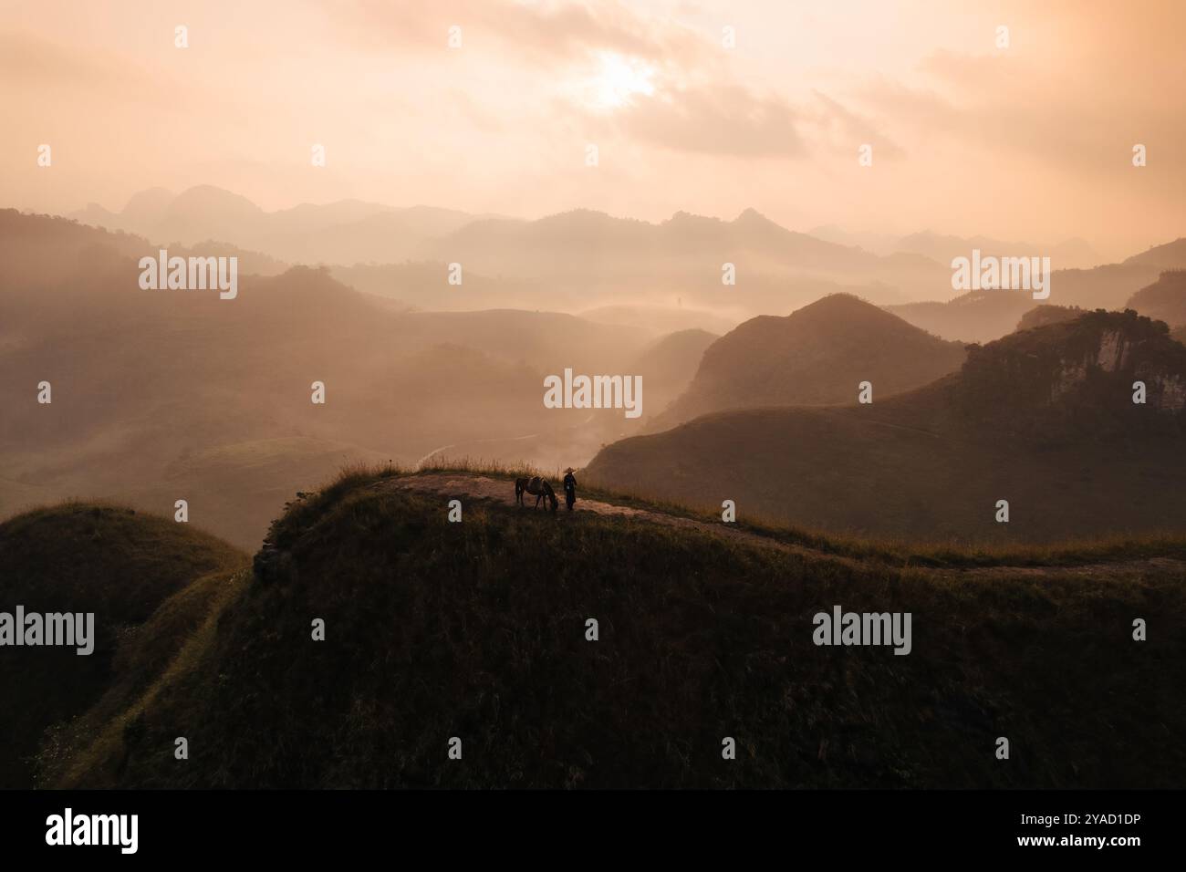 Blick aus der Vogelperspektive auf den wunderschönen Sonnenaufgang über Ba Guang Grashügel, Bergkette und Nebel zusammen mit Dorfbewohnern, die ihr Pferd auf den Berg bringen Stockfoto