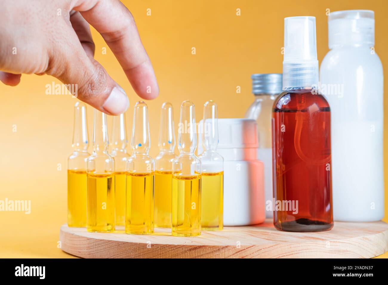 Eine Hand, die eine Ampulle hält, gefüllt mit gelber Flüssigkeit, umgeben von verschiedenen Flaschen mit Hautpflegeprodukten in verschiedenen Formen und Größen. Der Hintergrund Stockfoto